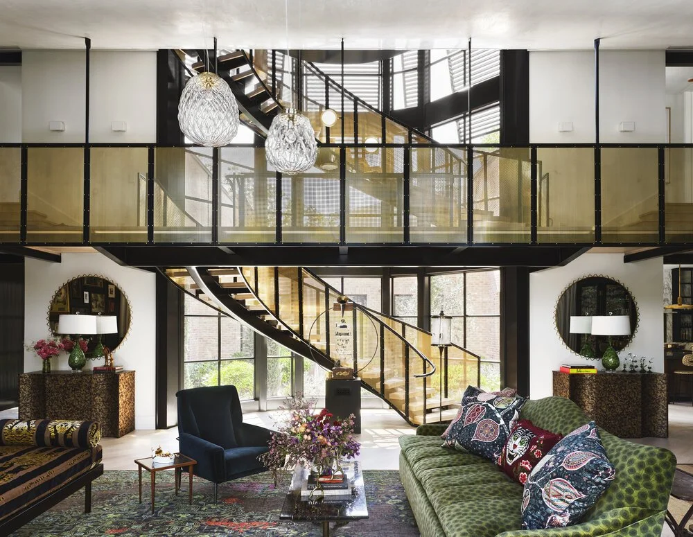 Modern living room with a stylish lofted design, featuring a green patterned sofa with decorative pillows, a blue armchair, floral decorations, and a spiral staircase leading to an upper level. Two large hanging light fixtures and a colorful rug comp