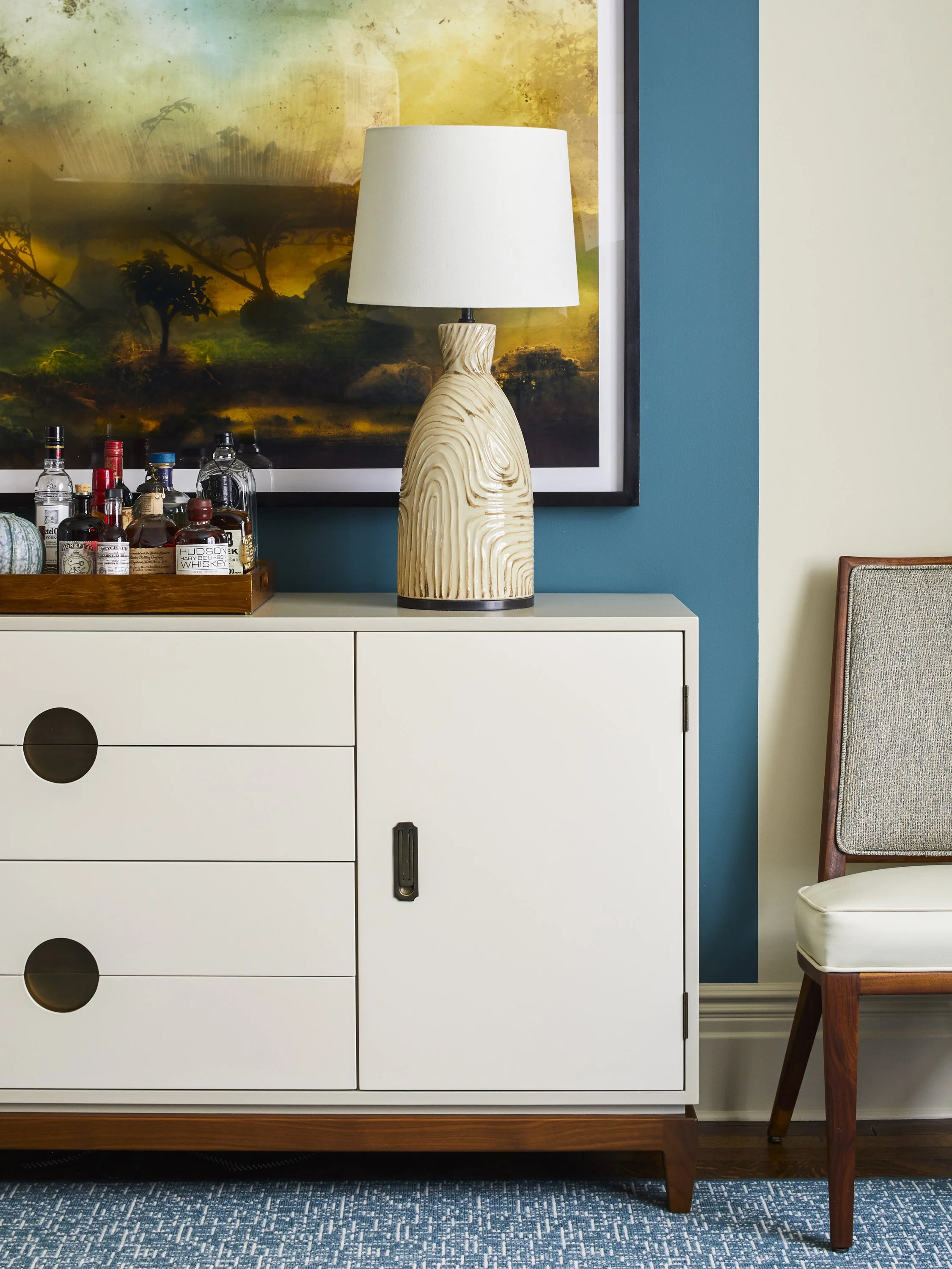 Interior of a room with a white cabinet featuring circular handles, topped with various liquor bottles on a tray and a ceramic table lamp with a white shade. The wall has a dark bluish accent color and a large framed artwork. A partial view of a chai