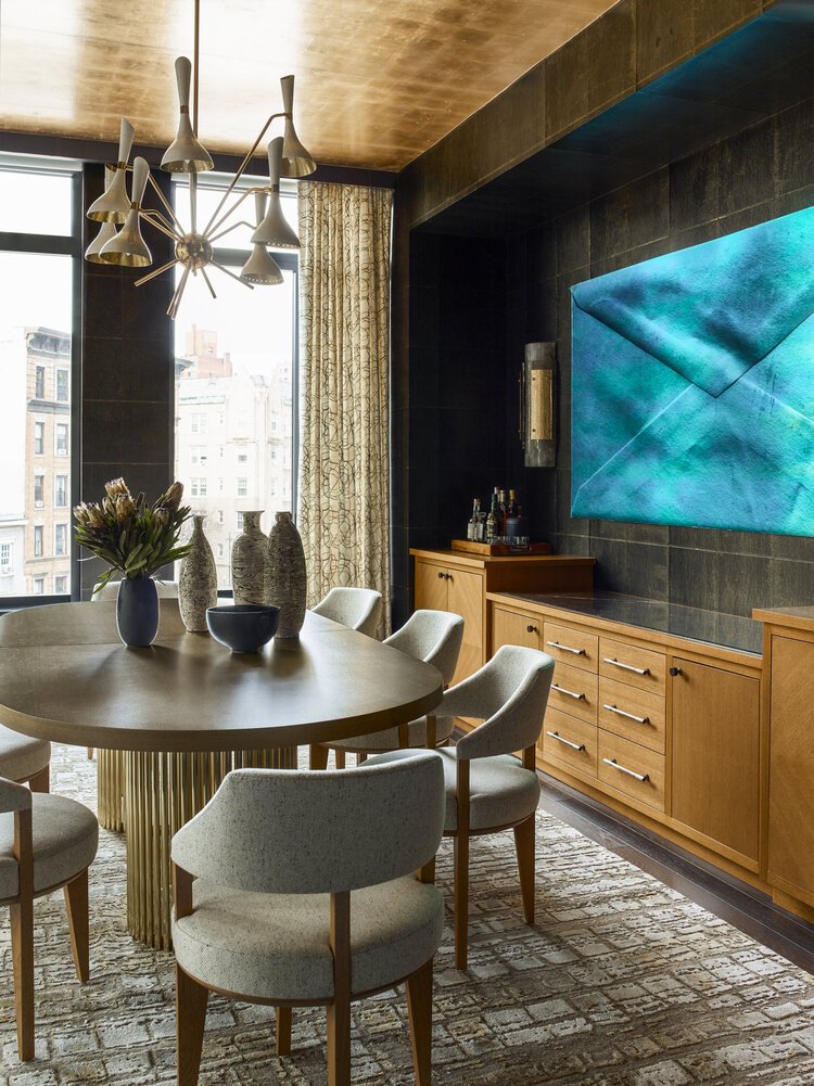 Modern dining room with a round wooden table, gray upholstered chairs, large abstract wall art, a decorative light fixture, and city view from the window.