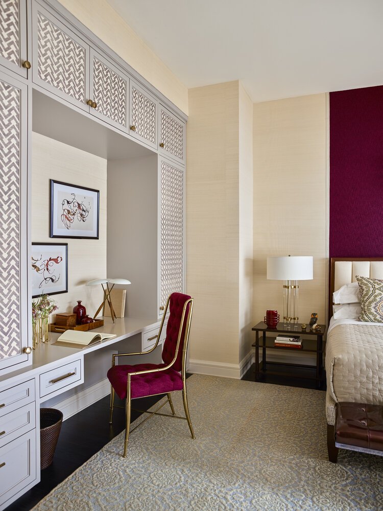 Elegant bedroom with a desk and chair. The desk is built into white cabinetry with patterned doors, adorned with decorative items and a lamp. A red accent chair is placed at the desk. The bed has gold and red accents with a nightstand, lamp, and deco