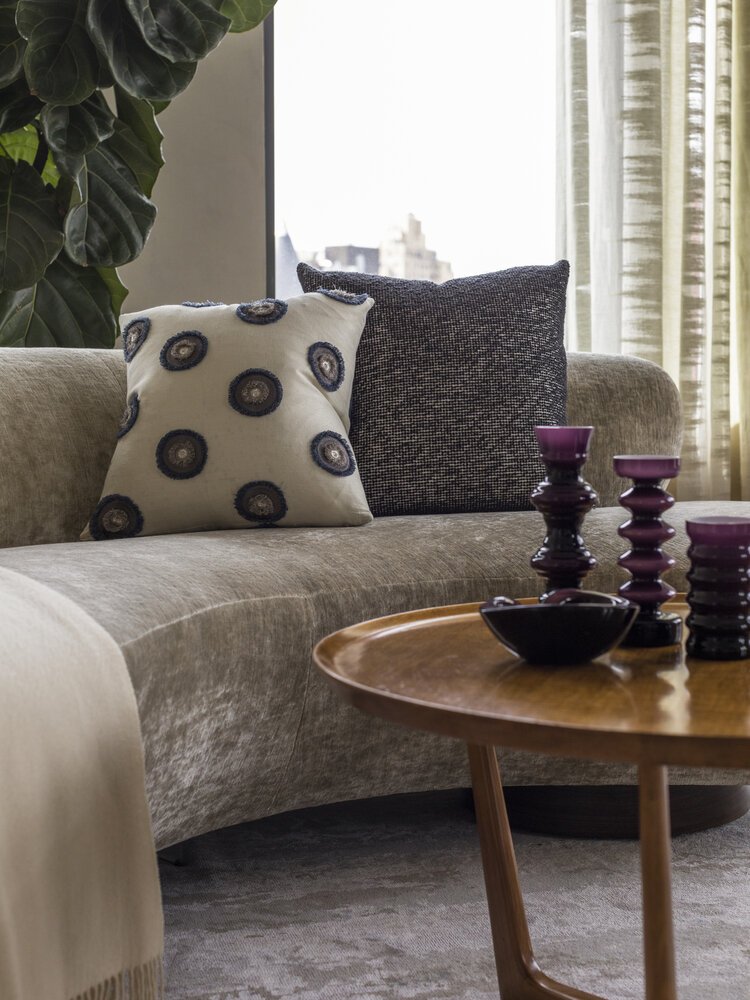 Modern living room with a curved beige sofa, two patterned throw pillows, a wooden coffee table, purple decorative items, and large green plant.