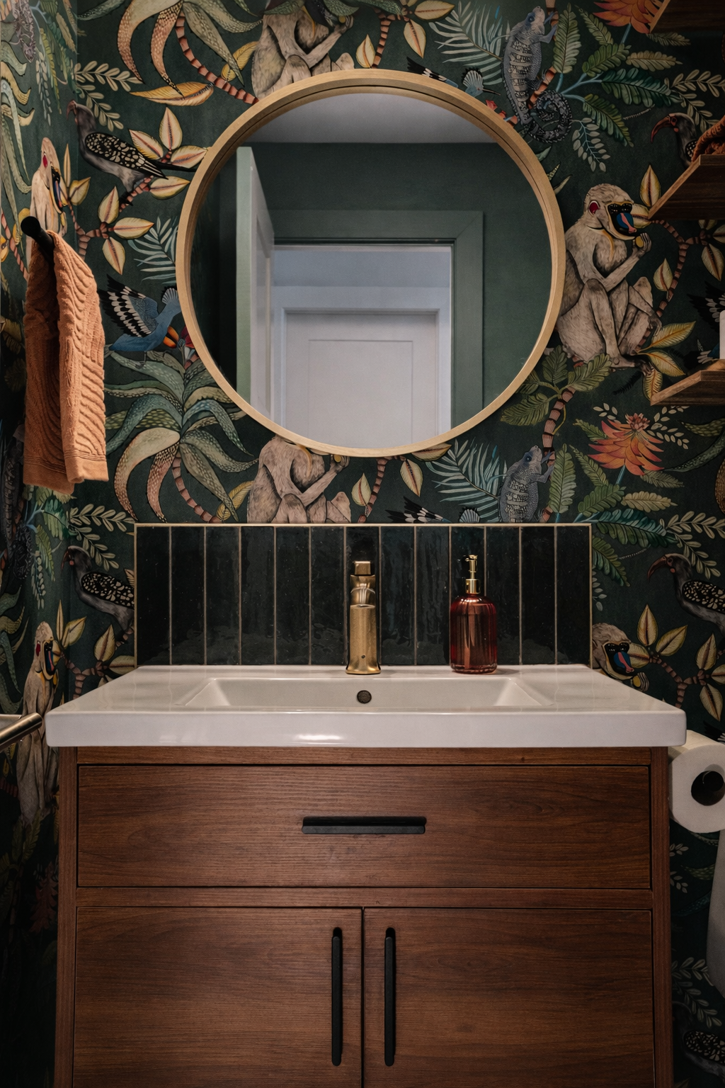 A bathroom with jungle-themed wallpaper featuring animals such as monkeys, birds, and chameleons. A round mirror is mounted above a white sink with a gold faucet and a red soap dispenser. There is a wooden vanity beneath the sink, a towel hanging on 