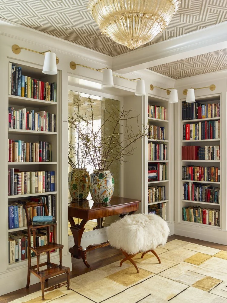 A luxurious home library with built-in bookshelves filled with books. A wooden side table with two decorative vases and branches stands against a mirrored wall. A fur-covered stool is positioned nearby. The ceiling features a geometric pattern and an