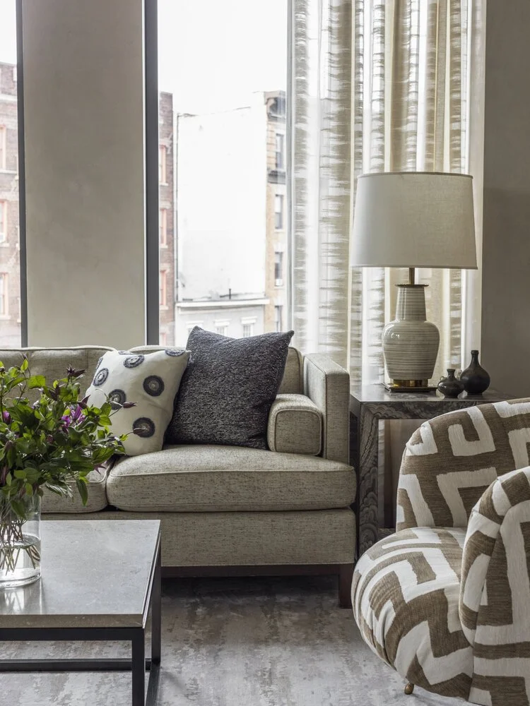 Modern living room with a beige armchair and patterned cushions, a side table with a ceramic table lamp, geometric-patterned chair, large window with curtains, and a coffee table with a vase of flowers.
