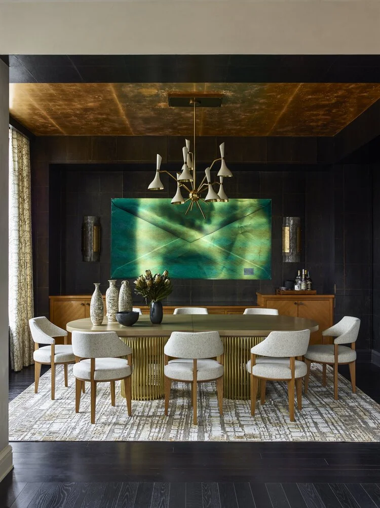 Modern dining room with an oval table surrounded by white cushioned chairs, a large abstract painting in green tones on the wall, decorative vases on the table, a contemporary light fixture above, and a patterned rug on dark wood flooring.