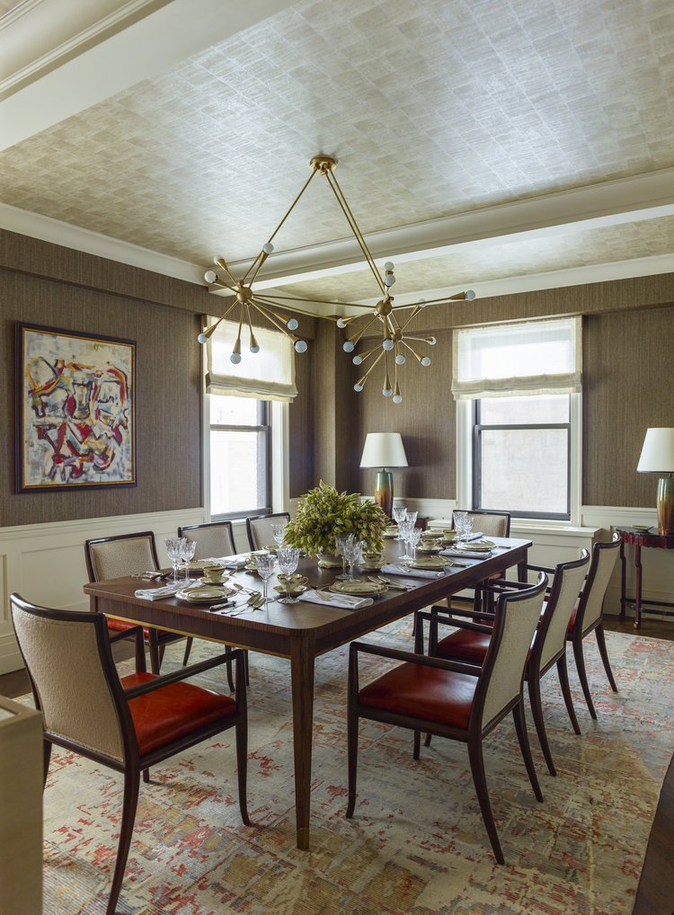 Elegant dining room with a modern chandelier, wooden dining table set for eight, upholstered chairs, large windows with light fixtures, and a colorful abstract painting on the wall.
