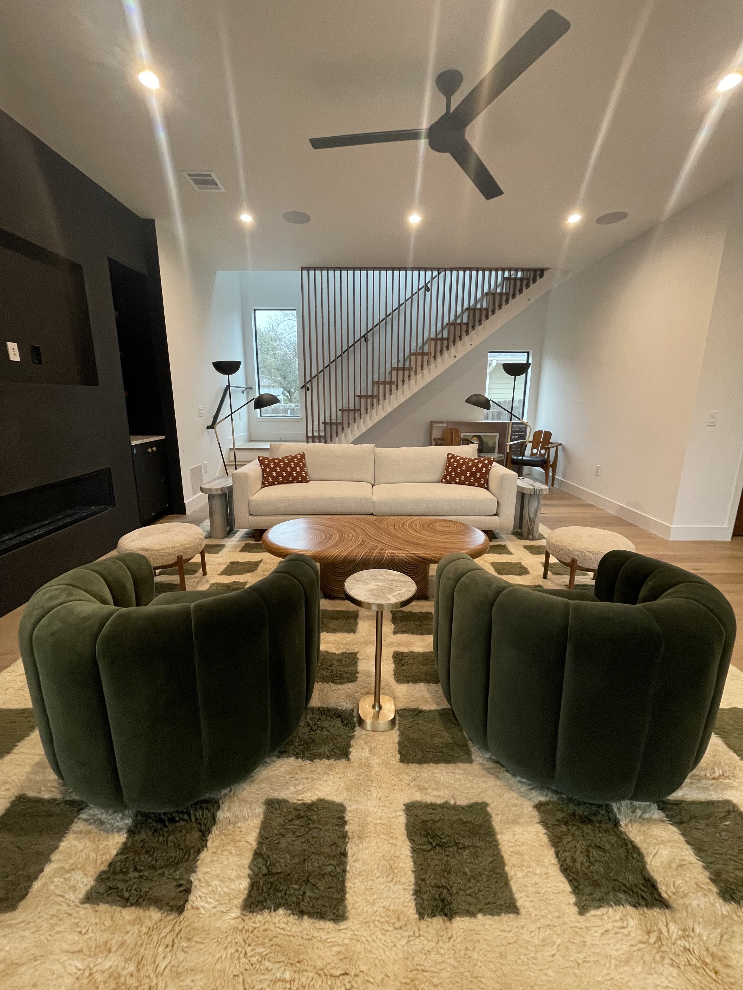 Modern living room with dark green velvet chairs, round wooden coffee table, cream sofa, geometric carpet, floor lamps, and a staircase with wooden slats in the background.