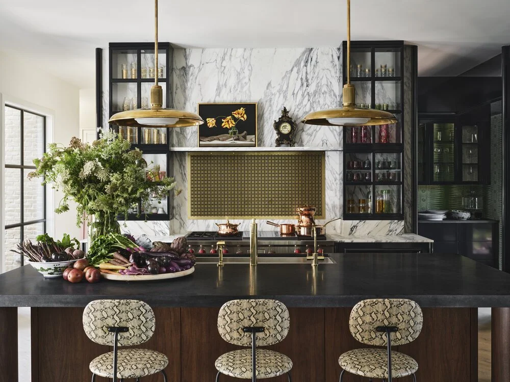 Modern kitchen with marble backsplash, gold accents, and decorative shelves. Large island with snake-patterned stools. Fresh vegetables and flowers displayed on countertop.
