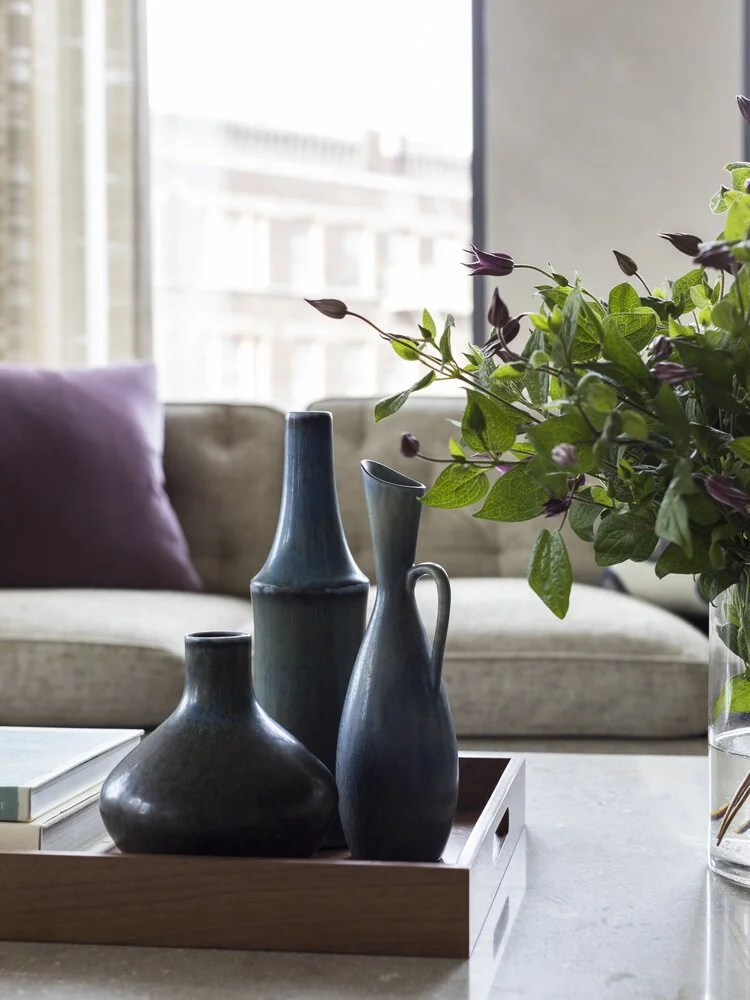 Decorative vases on a wooden tray with green leaves in a vase, sofa in the background.