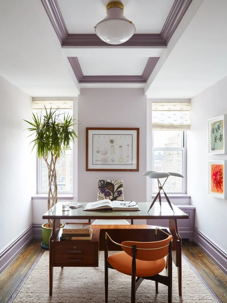 Modern home office with wooden desk, orange chair, framed art, large window, hanging light fixture, and indoor plant.