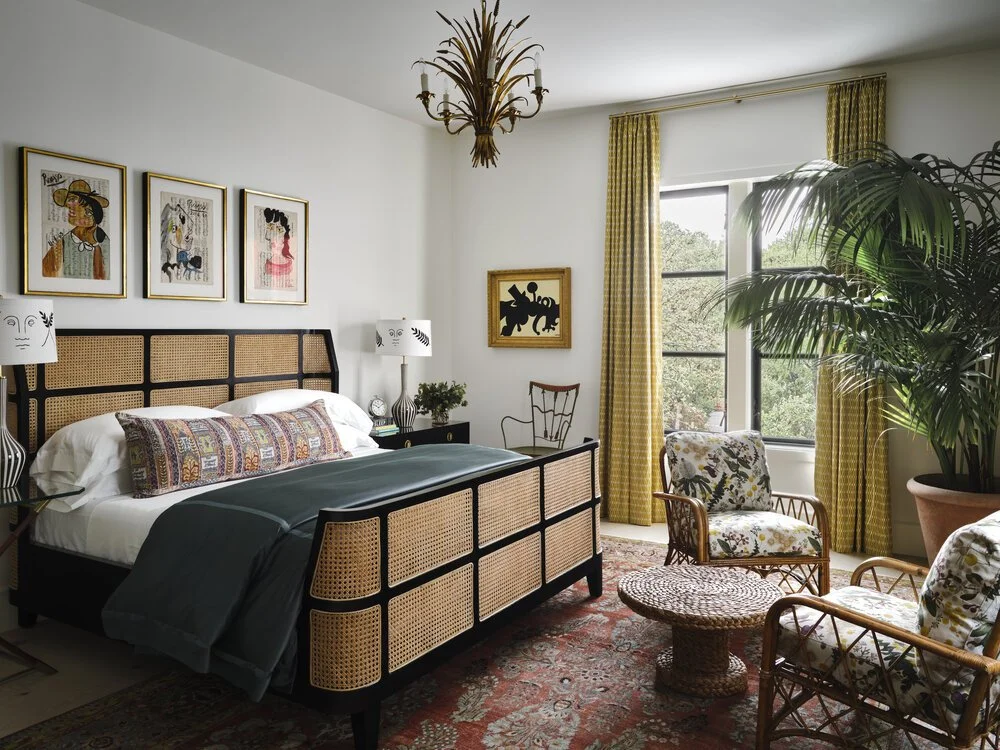 A stylish bedroom with a wicker bed, patterned cushions, and a rattan coffee table. The room features an eclectic mix of framed art on the walls, green bedside lamps, large yellow curtains, and a lush potted plant. A colorful rug covers the wooden fl
