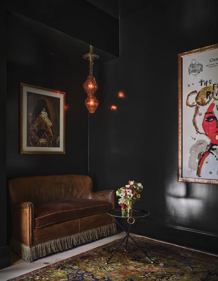 A small, stylish corner with dark walls featuring a brown leather loveseat with fringe at the bottom. Above it hangs an ornate pendant light. The walls display two framed artworks: one on the left showing a historical figure, and one on the right wit