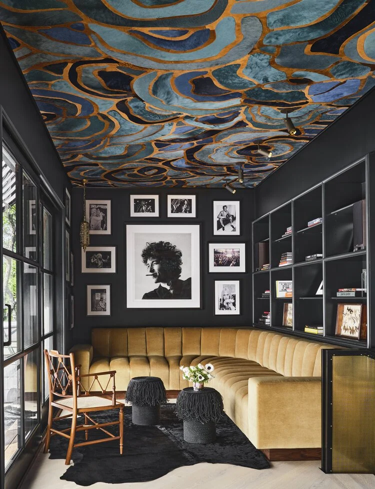 Stylish living room with abstract ceiling design, black walls, and framed photos. Features a mustard yellow sectional sofa, dark shag rug, round fringe-topped stools, wooden chair, and wall shelves.