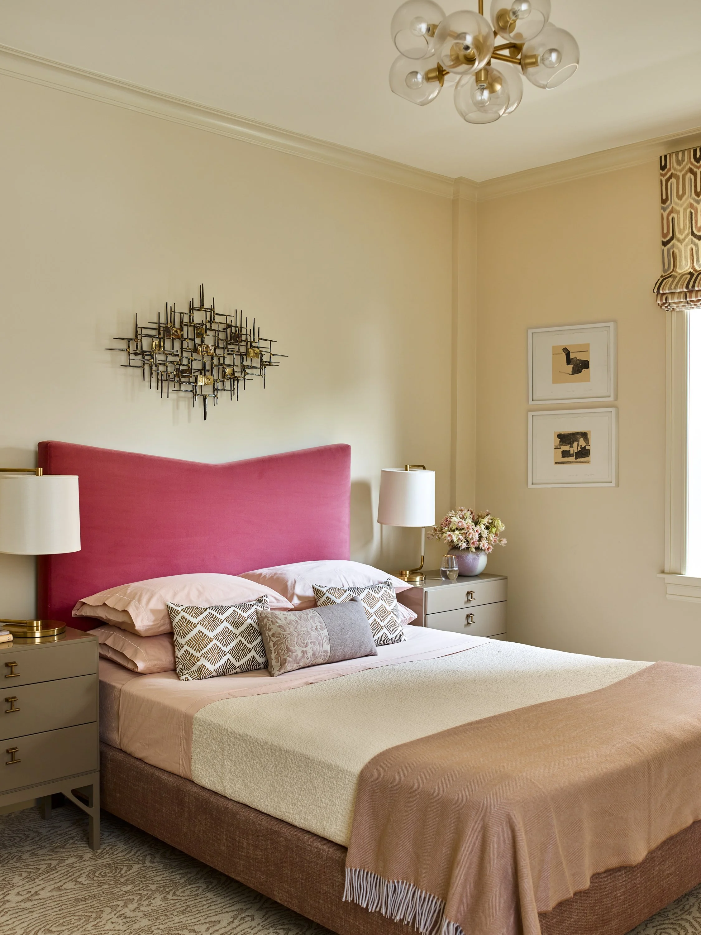 Bedroom with a pink upholstered headboard, beige walls, and modern decor including a metal wall sculpture, dual bedside tables with lamps, and decorative pillows on the bed.