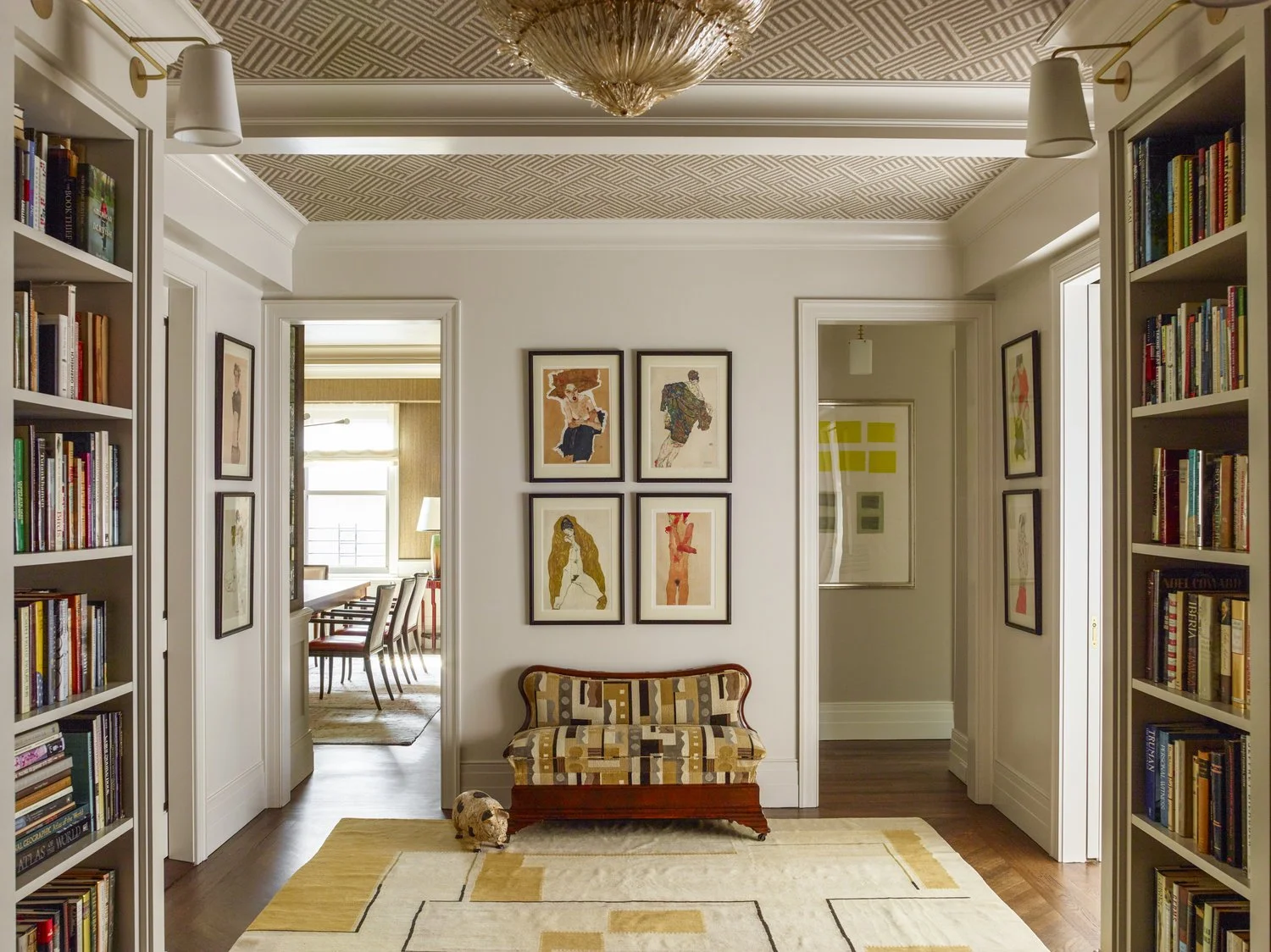 A stylish hallway with bookshelves on both sides, filled with books, leading to a dining area. The hallway has framed artwork on the walls, a small patterned bench, and a geometric-patterned ceiling. There is a patterned area rug on the wooden floor 