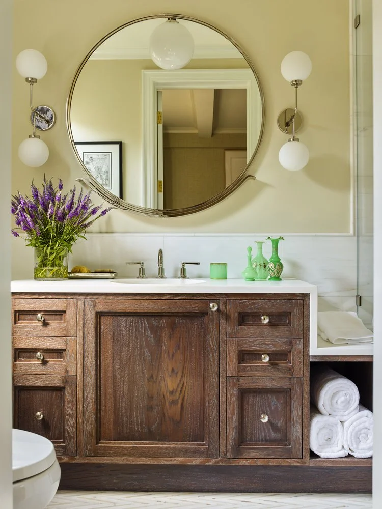 Bathroom vanity with large round mirror, wooden cabinet, elegant sconces, and rolled white towels. Lavender flowers in a vase and decorative bottles are on the countertop.