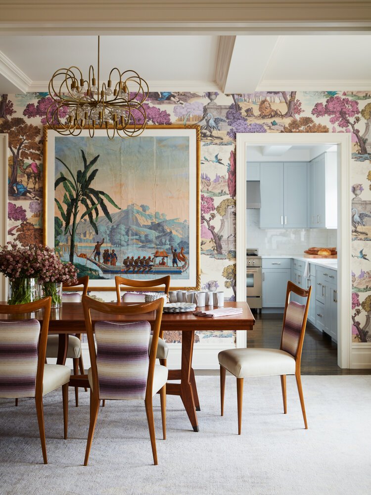 Dining room featuring a wooden table with upholstered chairs, a floral wallpaper, a large framed artwork with a tropical scene, and a chandelier. A view of the adjacent modern kitchen is visible through a doorway.