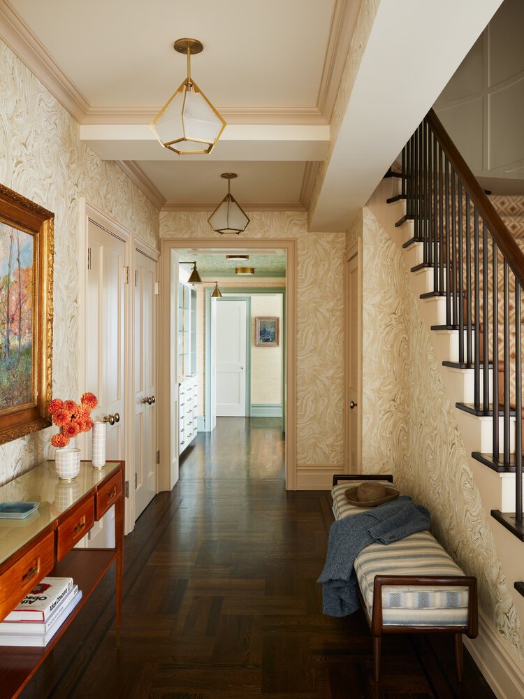 Elegant hallway with patterned wallpaper, wooden floors, and a staircase. Features a wooden table with vases and books, a striped bench with a hat and sweater, geometric pendant lights, and artwork on the walls.
