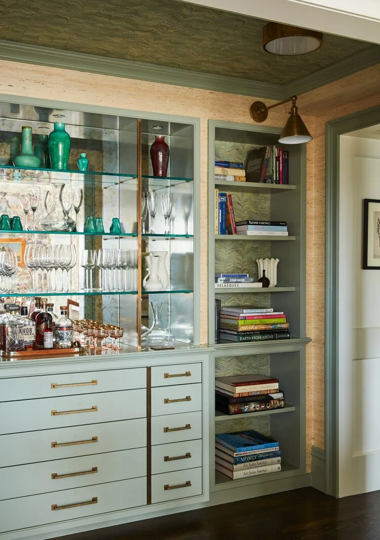 Interior with glass cabinet displaying vases and drinkware, alongside shelves with books and a light green cabinet with drawers.