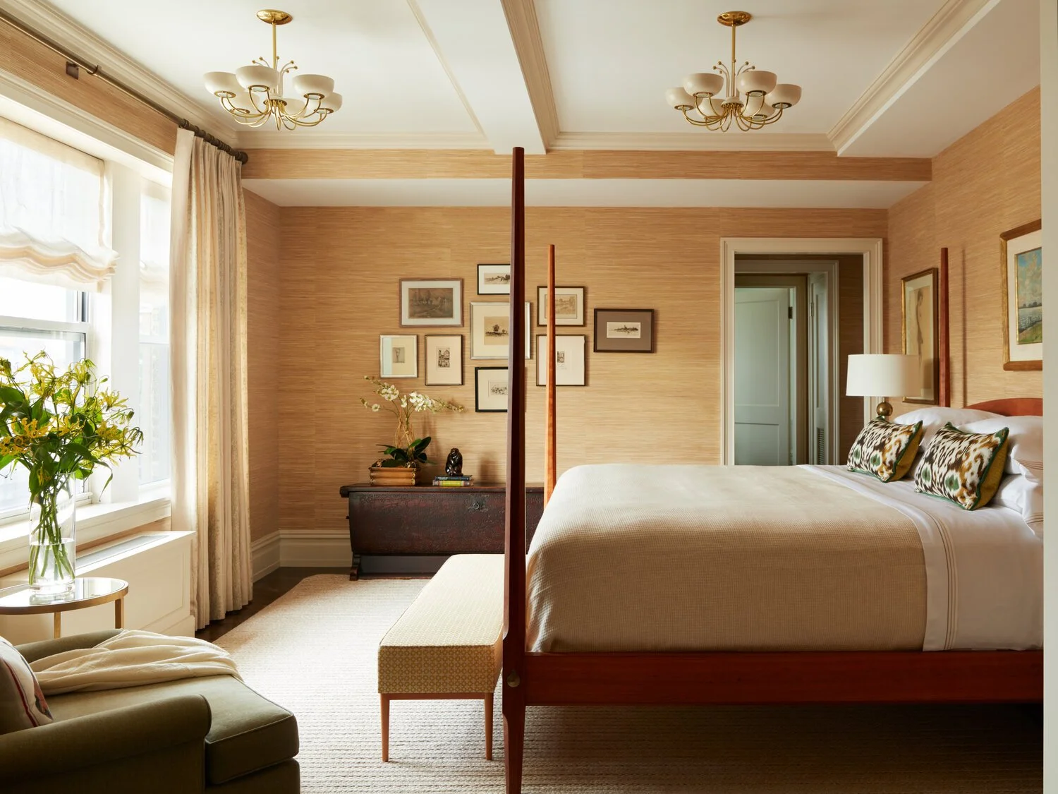 Elegant bedroom interior with beige walls and a large bed featuring patterned pillows. The room has a bench at the foot of the bed and a comfortable armchair with a throw on it. Large windows are adorned with light curtains. The walls are decorated w