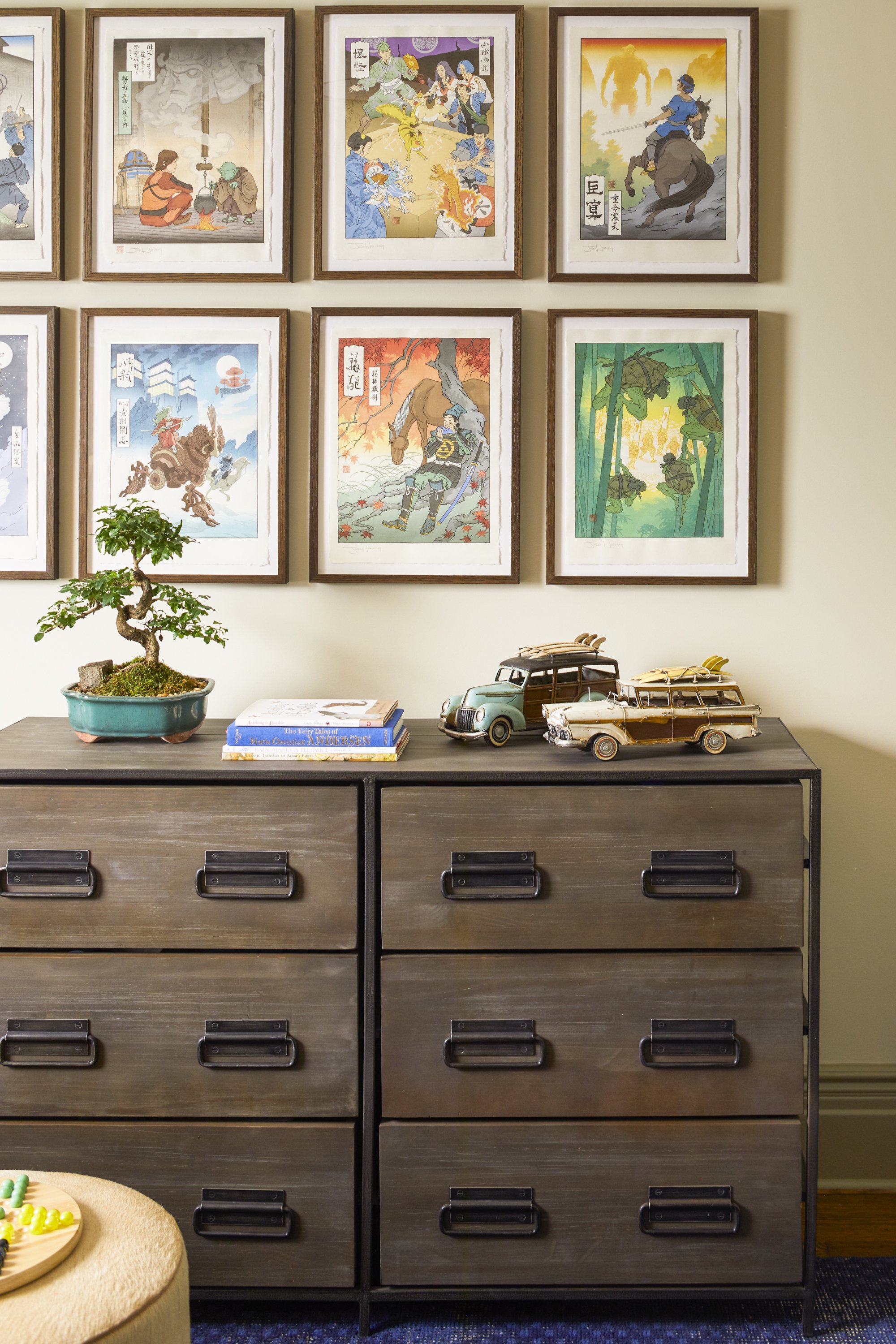 A wooden dresser with six drawers, featuring two vintage toy cars and two books on top, alongside a small potted bonsai tree. Above the dresser, there are nine framed, colorful art prints in a grid pattern on the wall.