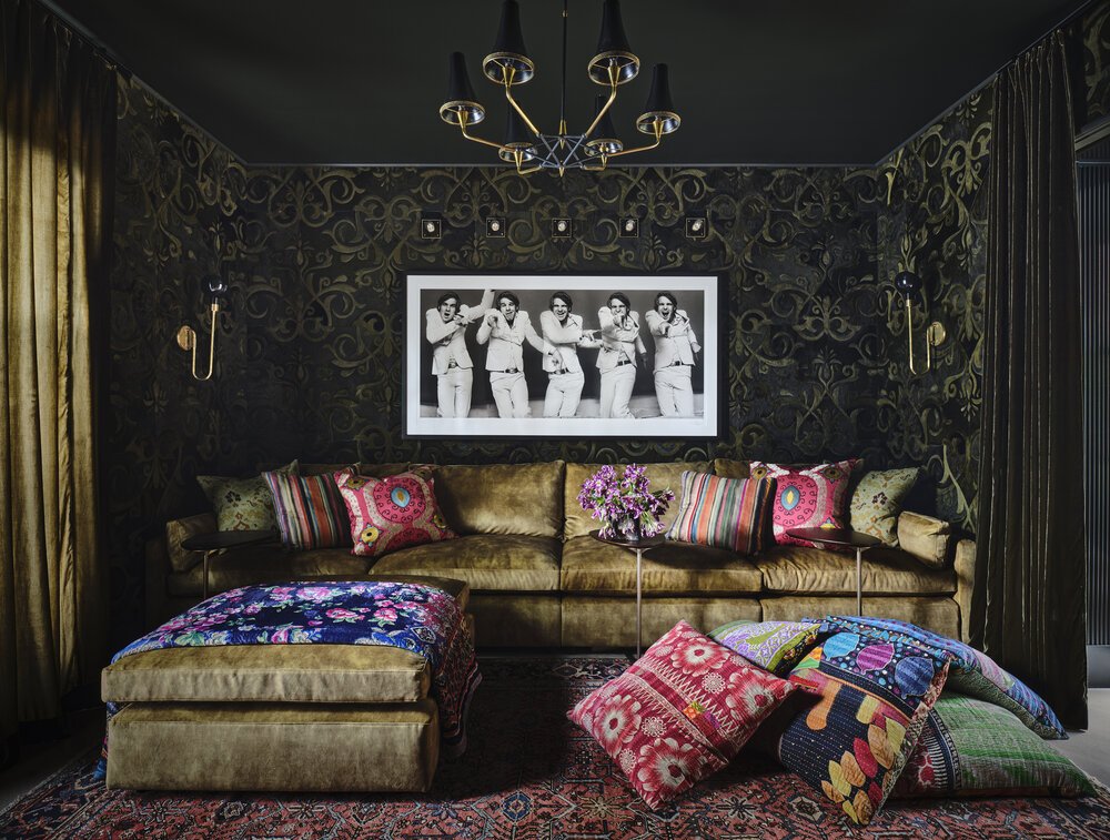 Luxurious living room with dark patterned walls, a large velvet sofa adorned with colorful pillows, a matching ottoman, floral arrangement on a small table, and a black-and-white photograph framed on the wall.