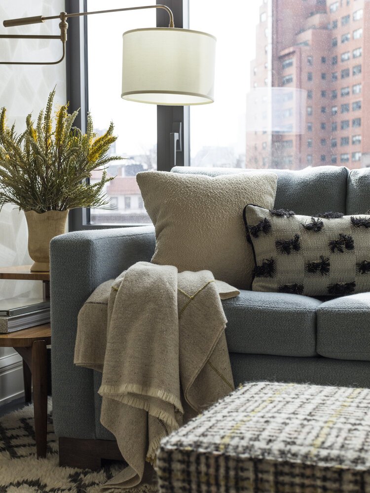 Cozy living room with blue sofa, patterned cushions, beige throw, side table, potted plant, and floor lamp, in front of large window with city view.