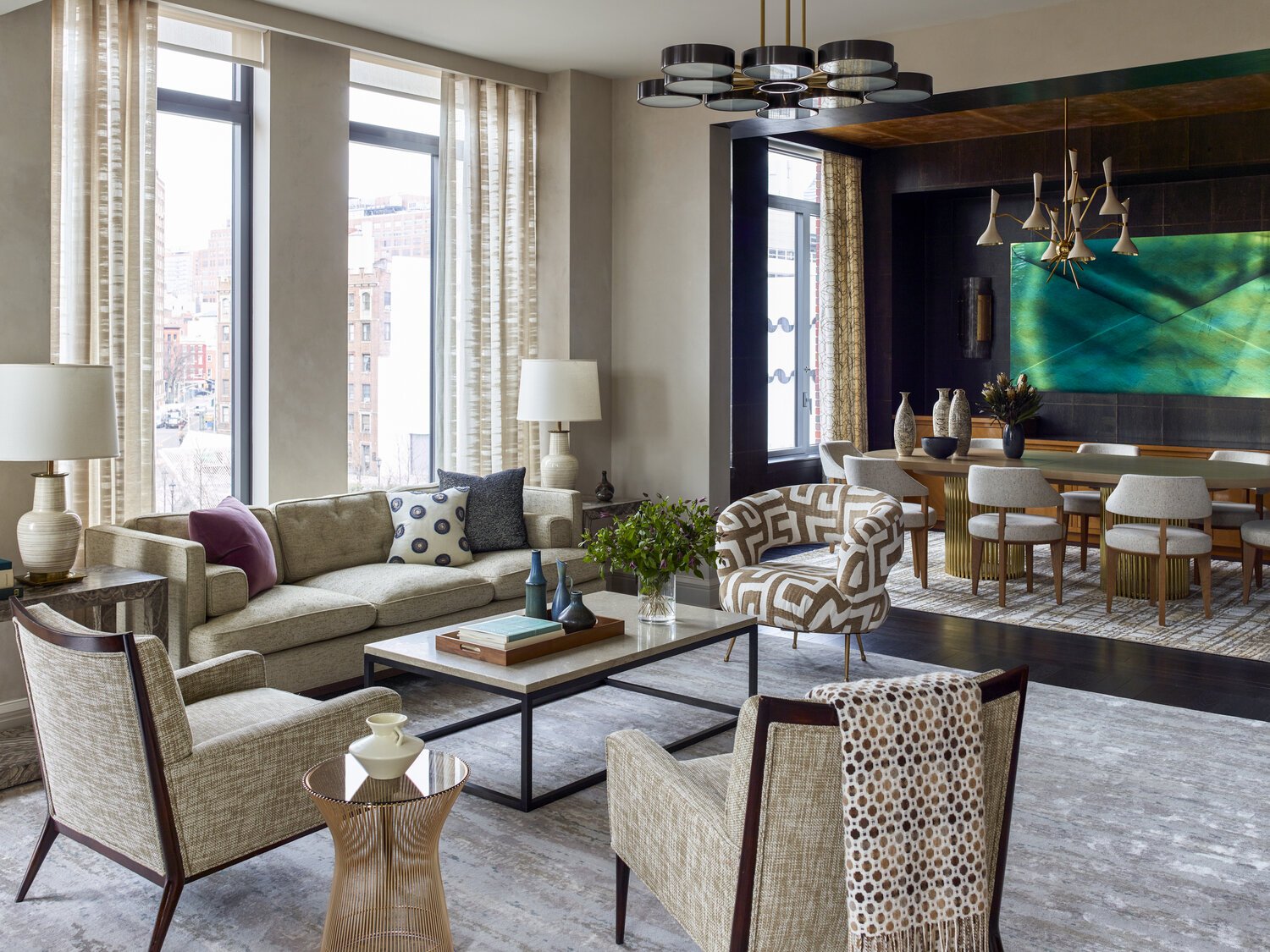 Modern living and dining room with large windows, beige sofa, patterned armchair, coffee table, abstract painting, and decorative lighting.