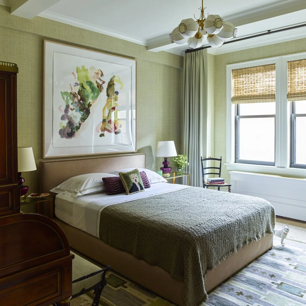 Contemporary bedroom with a large painting above the bed, neutral tones, textured bedding, two table lamps, and a decorative chair by a window with woven blinds.