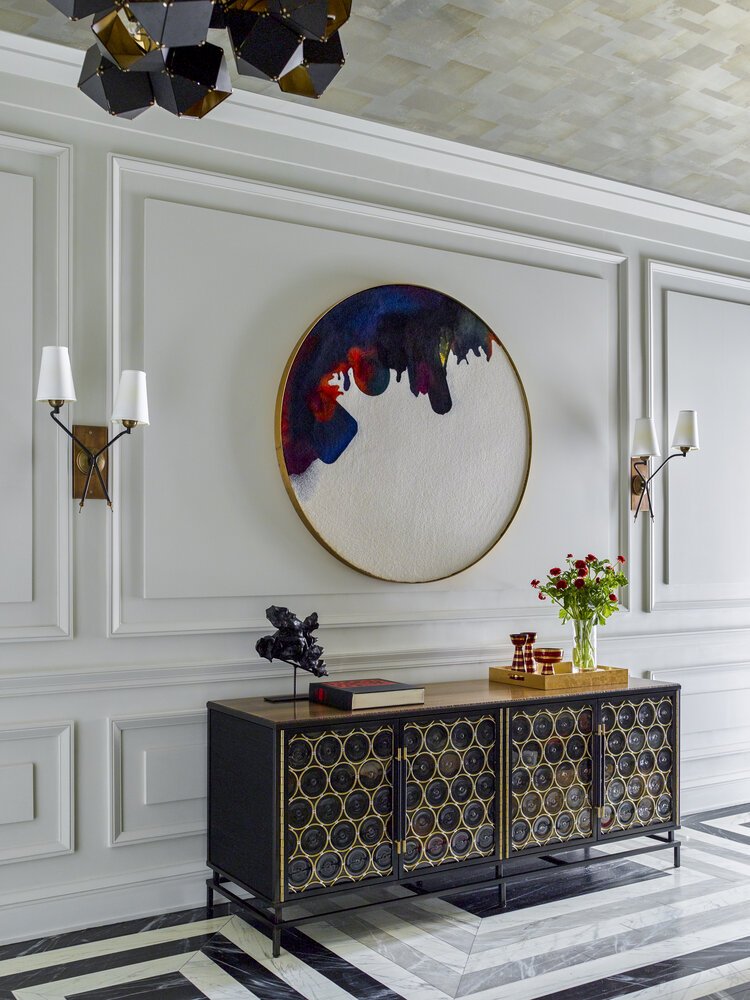 Modern interior with a black decorative cabinet featuring circular patterns, a colorful abstract circular wall art, two wall sconces with white shades, and a geometric black chandelier. On the cabinet are a sculpture, a book, and a vase with red flow