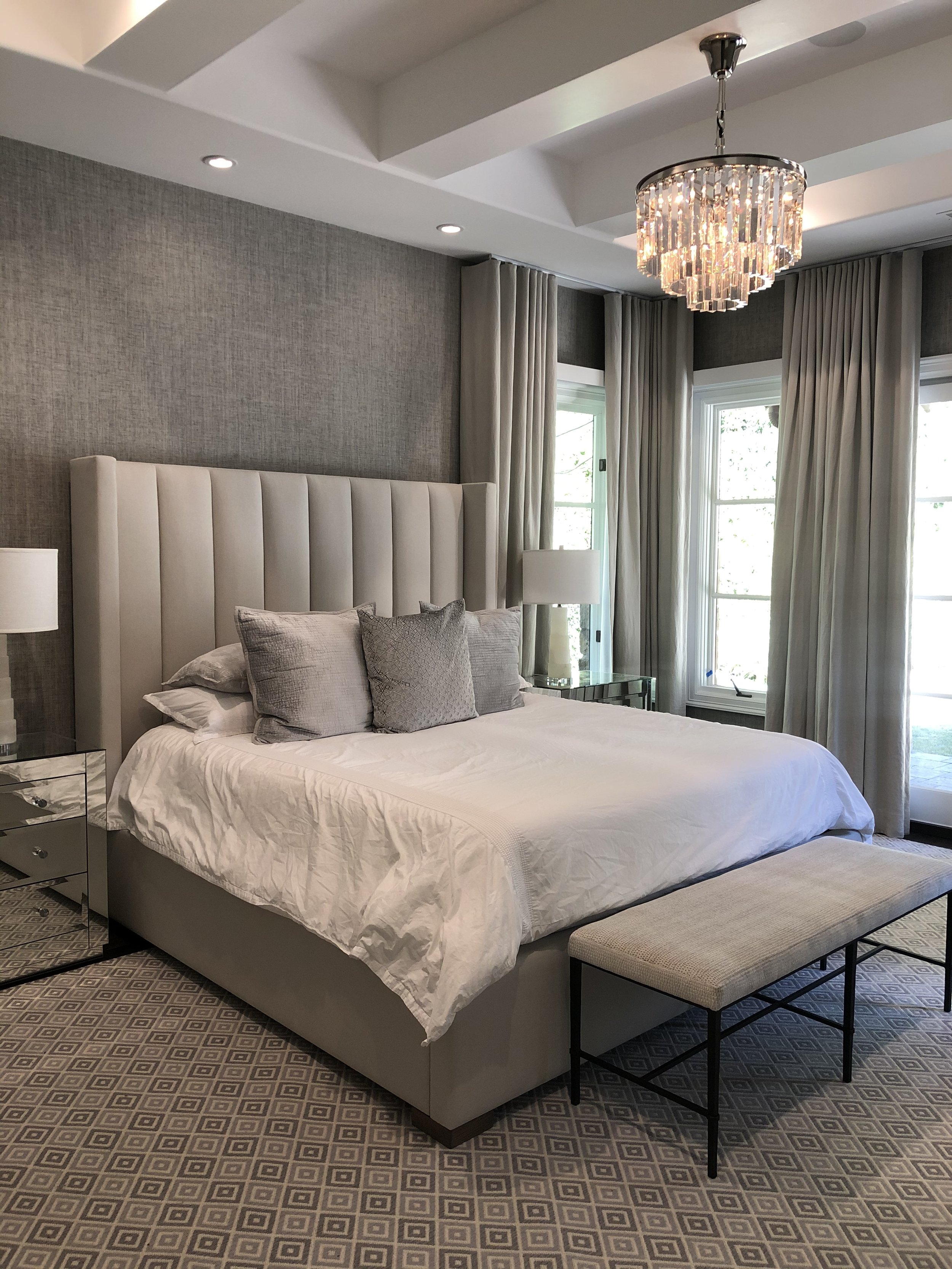 Elegant bedroom with a gray upholstered bed, white bedding, gray accent pillows, nightstands with lamps, a crystal chandelier, gray curtains, and a decorative carpet.