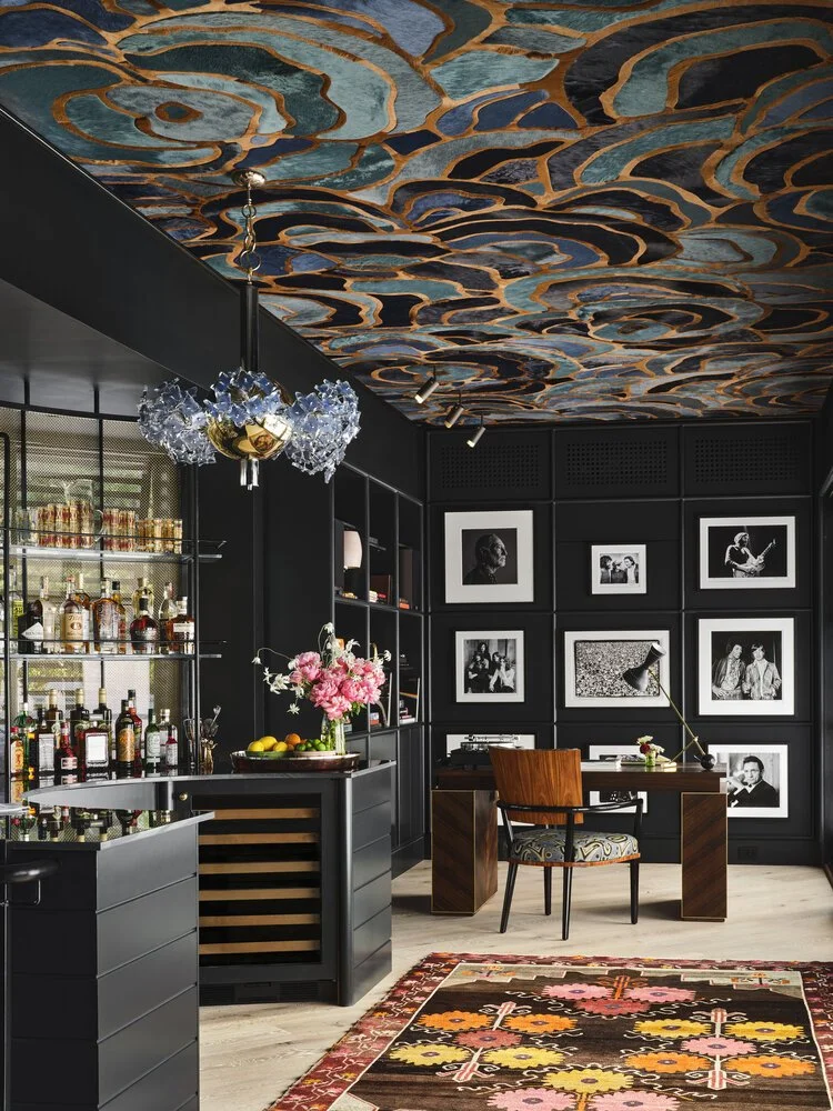 Luxurious home office with a modern bar area, featuring a colorful swirling ceiling pattern, black walls adorned with framed photographs, a sophisticated desk setup, stylish chandelier, and floral rug.