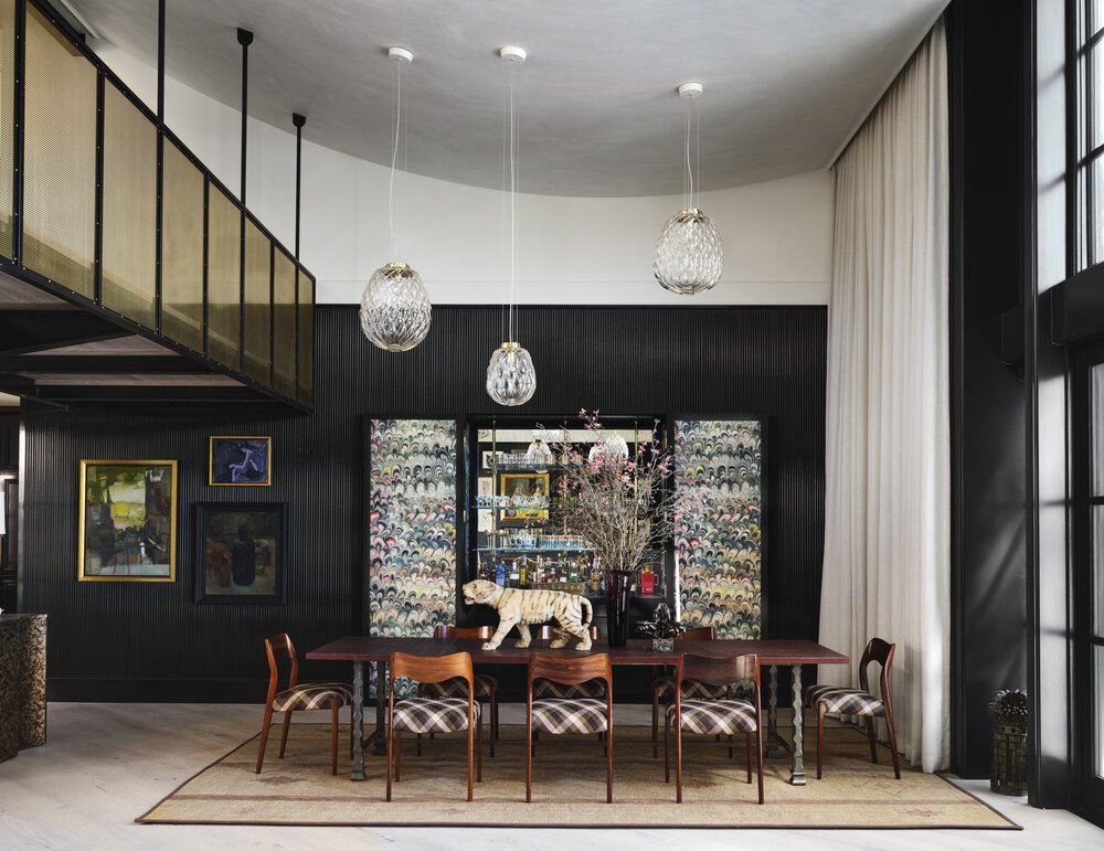 Modern dining room with a long wooden table and six chairs, featuring patterned upholstery. Glass pendant lights hang from the ceiling. A cabinet with colorful patterns and decorative pieces like a tiger statue and flower arrangement are in the backg