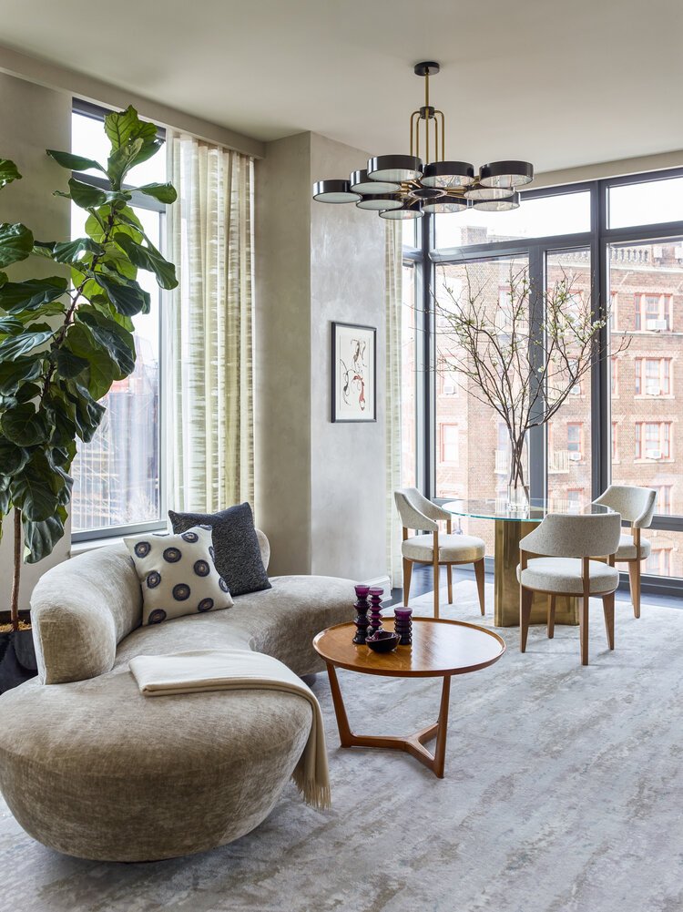 Modern living room with a round gray sofa, wooden coffee table, purple decor items, large plant, and a dining set near floor-to-ceiling windows. Overhead chandelier with black and gold accents. Natural light floods the room.