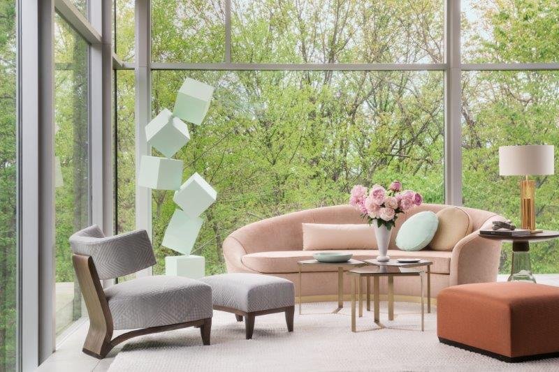 Modern living room with large windows, pink sofa, gray armchairs, and a vase of flowers on a round table. Outside, green trees are visible.