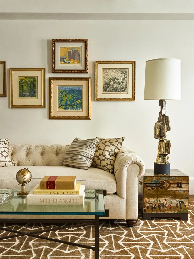 Elegant living room with a cream-colored sofa, geometric patterned pillows, decorative art on the wall, a modern table lamp, books and a small globe on a glass coffee table, and a patterned rug.