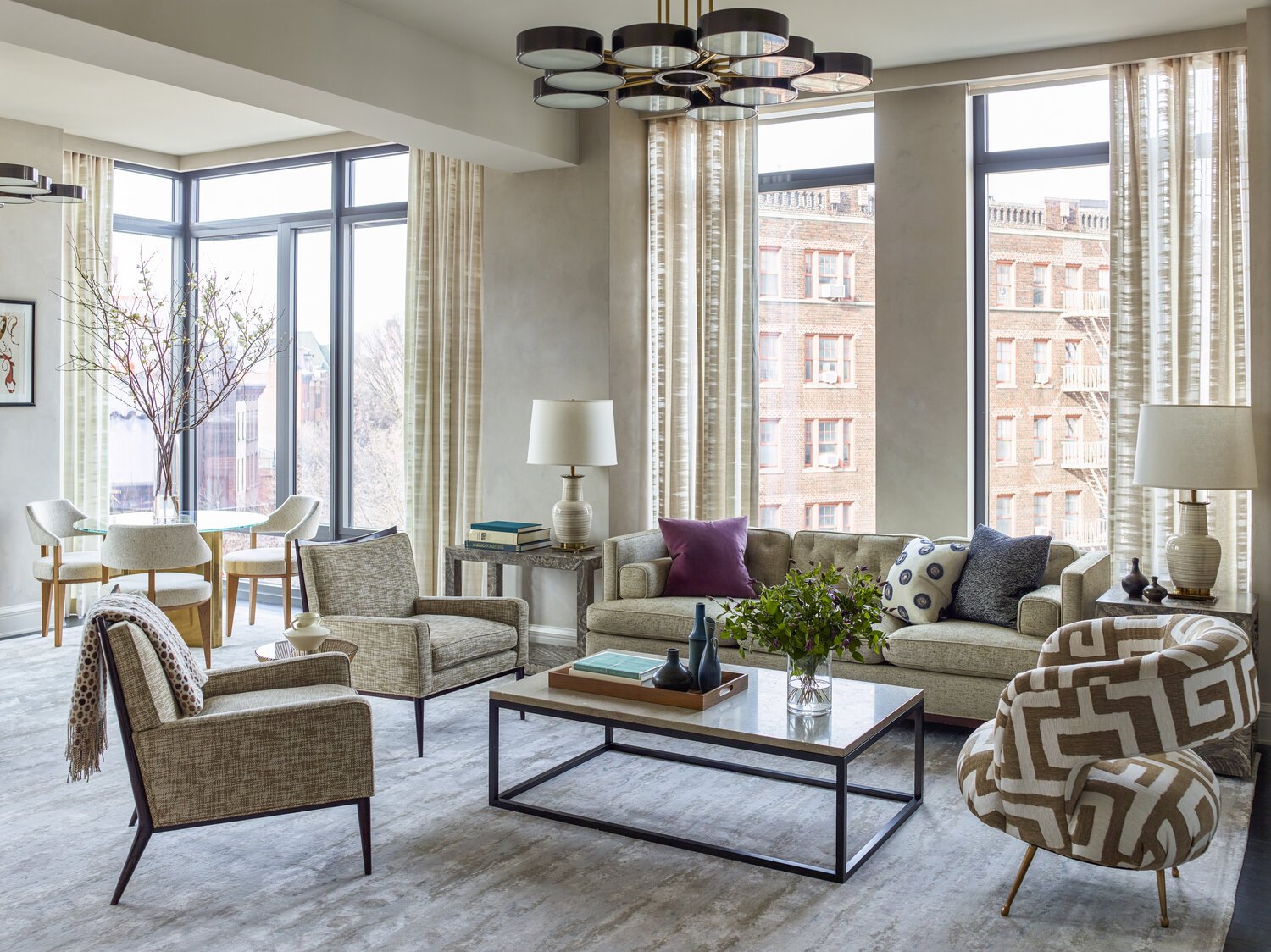 Modern living room with large windows, beige furniture, patterned armchair, coffee table, decorative vases, and potted plant, with cityscape view.