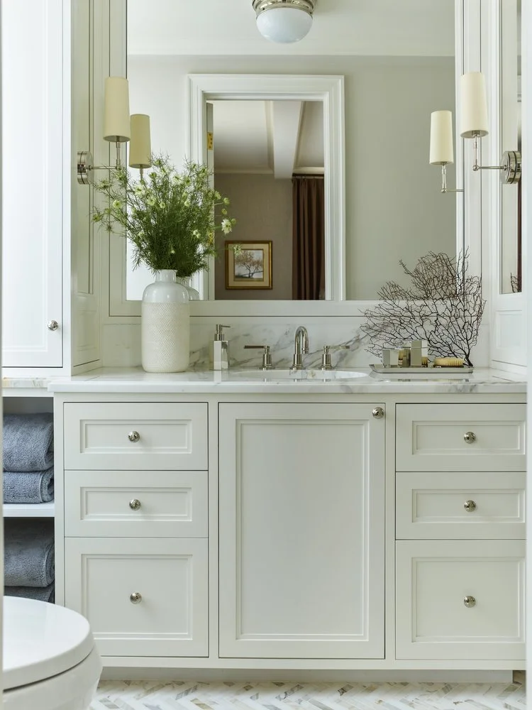 Elegant bathroom vanity with white cabinetry, marble countertop, central sink, and chrome faucet. Decor includes a vase with green plants, decorative branches, wall sconces, and a large mirror. Neatly folded gray towels are visible, and a ceiling lig