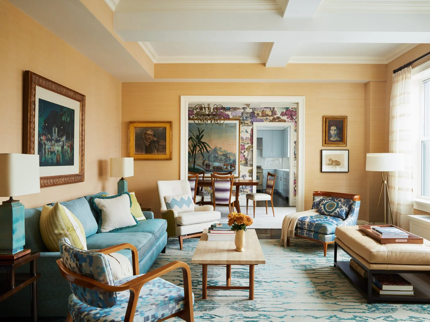 A stylish living room with a blue sofa, patterned chairs, and a coffee table. The walls are decorated with framed artwork, and there's a dining area visible through an open doorway. The room features a beige and blue color scheme, with a large window