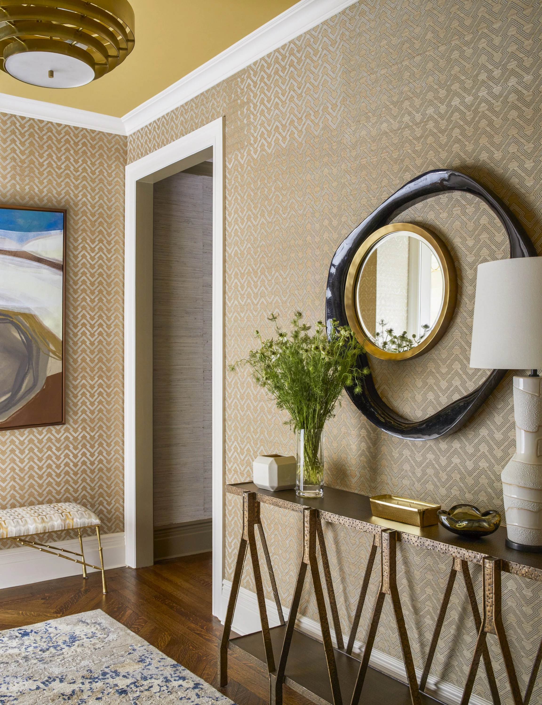 Interior room with patterned beige wallpaper, abstract art, gold ceiling light, a modern console table with decorative items, and a round mirror above it. A cushioned bench and a rug with blue and gray patterns are also visible.