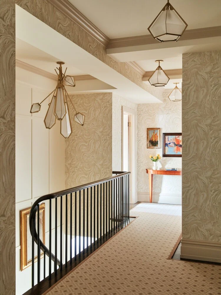 Elegant hallway with patterned wallpaper, beige carpet, modern geometric light fixtures, black railings, and framed artwork on the walls. A small console table with flowers and books is visible.