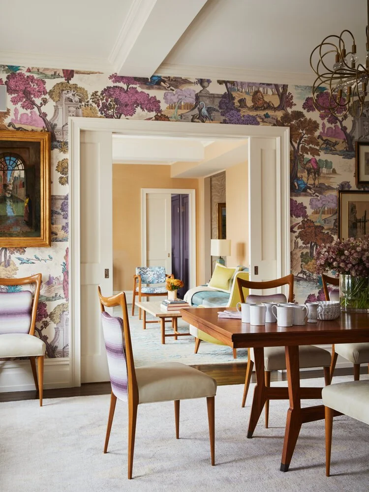 Elegant dining room with colorful wallpaper, wooden dining chairs, and a table set for tea. The room opens into a connected living area with a sofa, coffee table, and armchair, featuring a light color palette and textured walls.