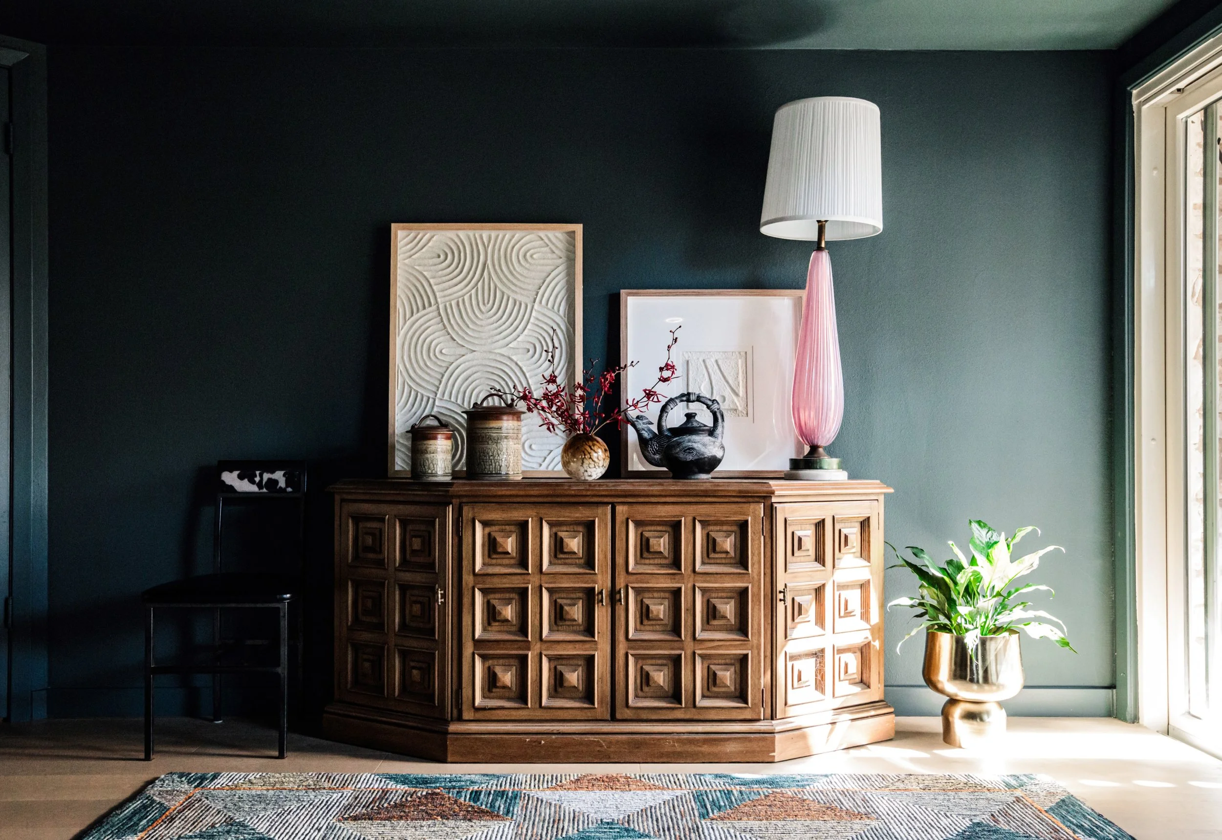Modern living room with a wooden sideboard, decorative vases, framed artwork, a tall pink lamp, patterned rug, and a potted plant.