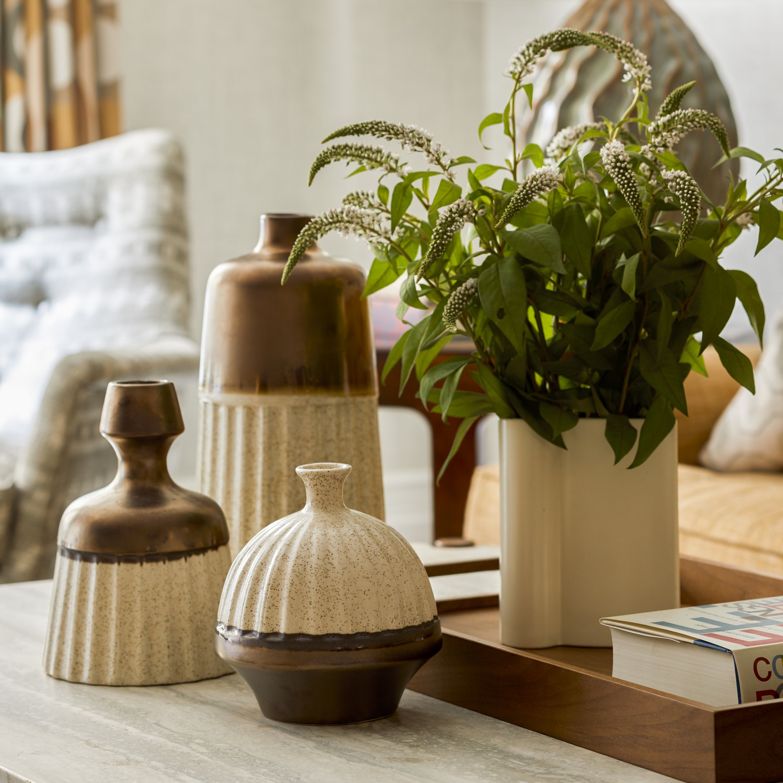 A living room table display with ceramic vases, a white rectangular vase with green leafy plants and white flowers, and a hardcover book on a wooden tray.