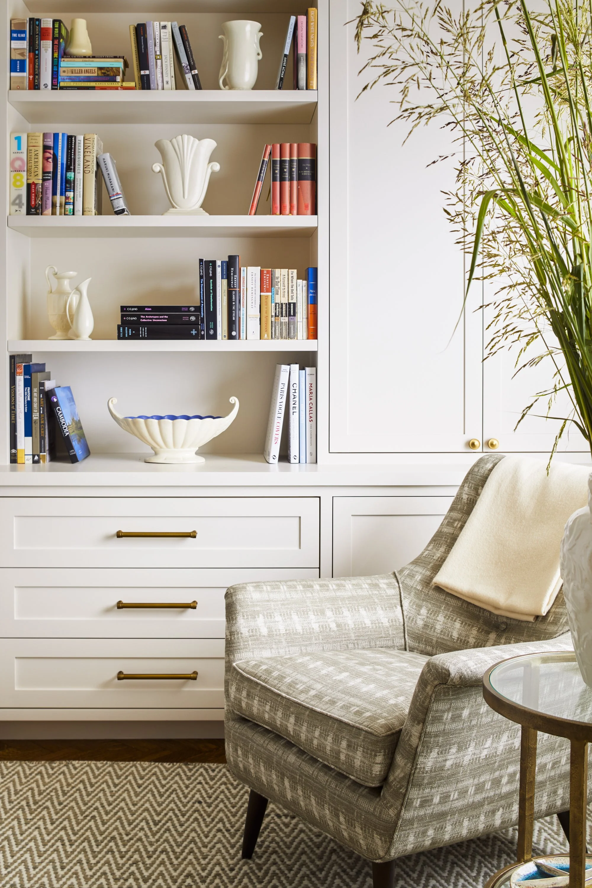 Cozy living room with bookshelves, a gray patterned armchair, elegant vases, and a large leafy plant. Books neatly arranged on shelves. White cabinets with brass handles enhance the decor.