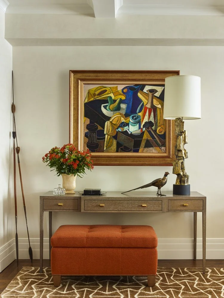 Room interior with a wooden console table, colorful abstract painting, decorative lamp, bird sculpture, floral arrangement, orange bench, and patterned rug.