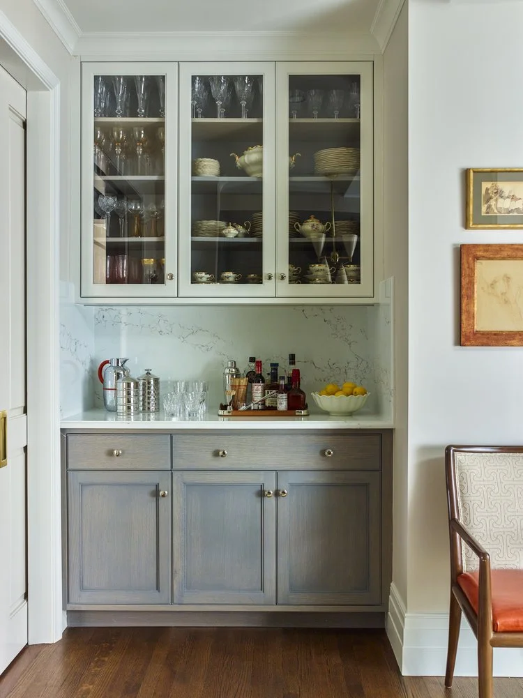 Elegant home bar setup with glass cabinet, wooden storage, and countertop featuring glassware, wine bottles, and a bowl of lemons.