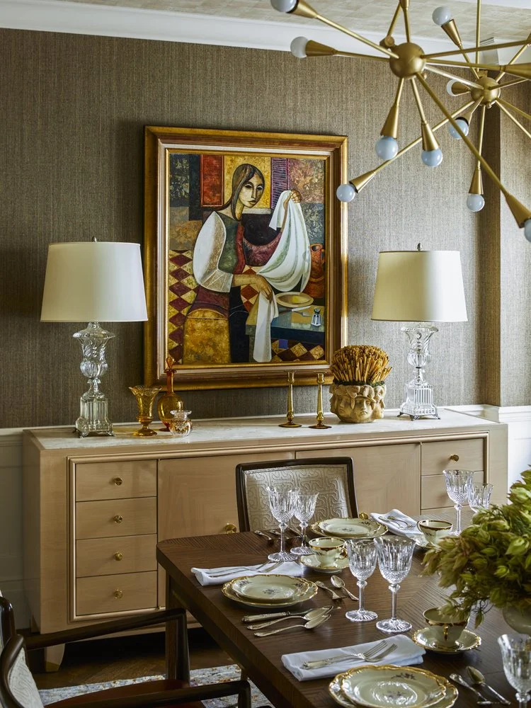 Elegant dining room with a wooden table set with fine china and crystal glasses. A sideboard topped with two ornate lamps and a framed painting hangs above. A modern chandelier decorates the ceiling.