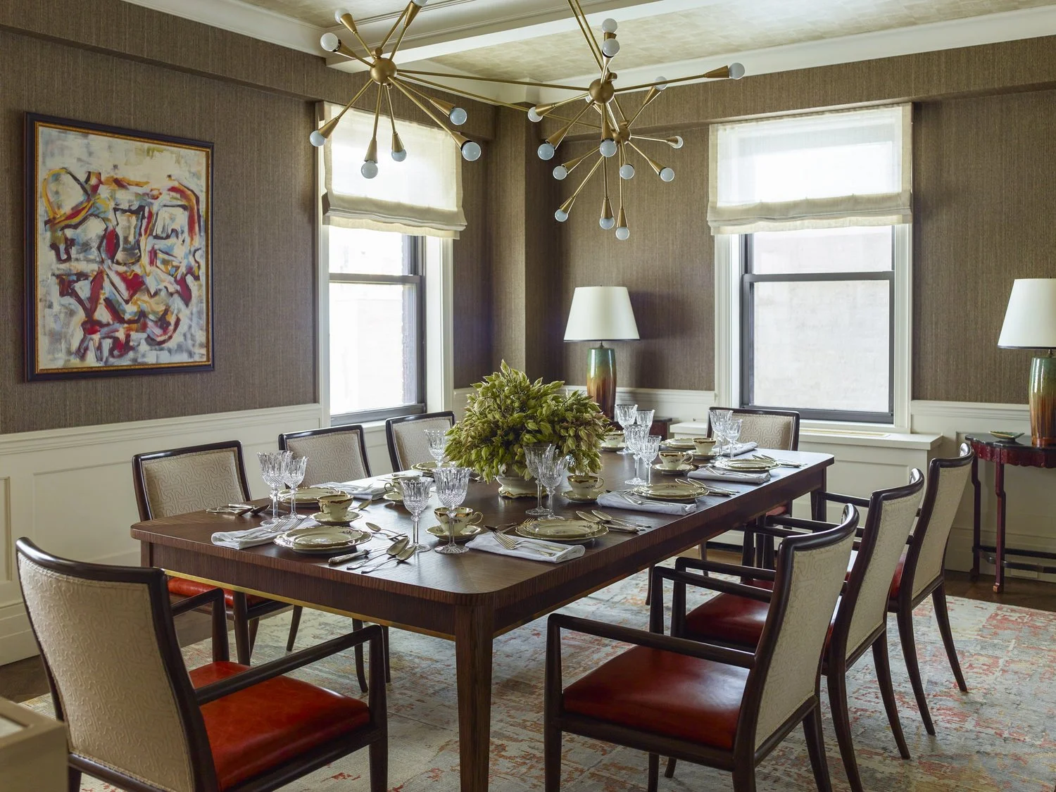 Elegant dining room with modern chandelier, rectangular dining table set for a meal, six upholstered chairs, abstract wall art, two table lamps, and large windows with Roman shades.