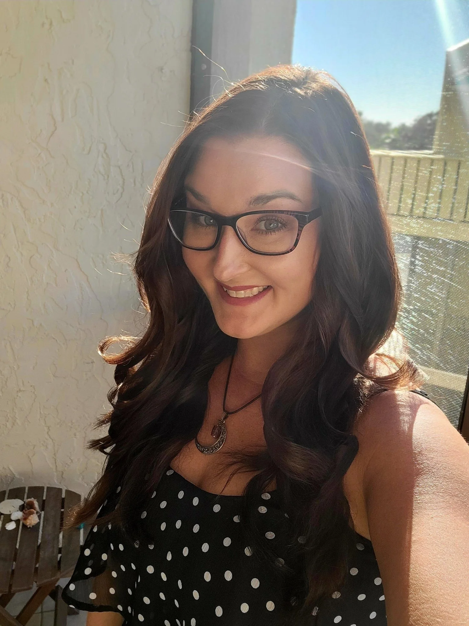 A woman with long wavy dark hair, wearing glasses, a black polka dot sleeveless top, and a necklace, smiling and taking a selfie in natural sunlight near a window.
